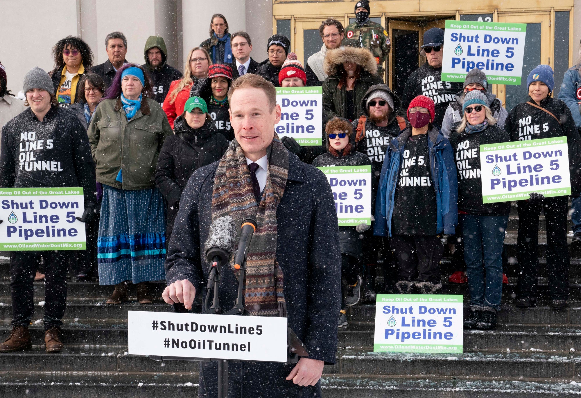 A man speaks at a podium with a crowd behind him, standing on steps. It is snowing. There are a lot of signs being held that read "Shut Down Line 5 Pipeline"