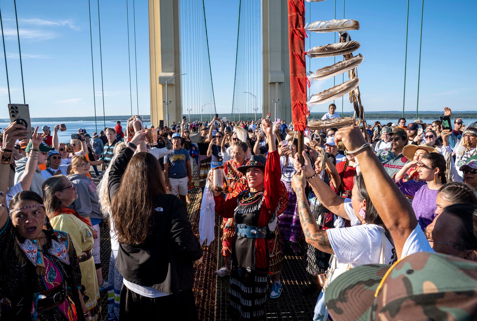 A large group of people stand on a suspension bridge over a lake and cheer.