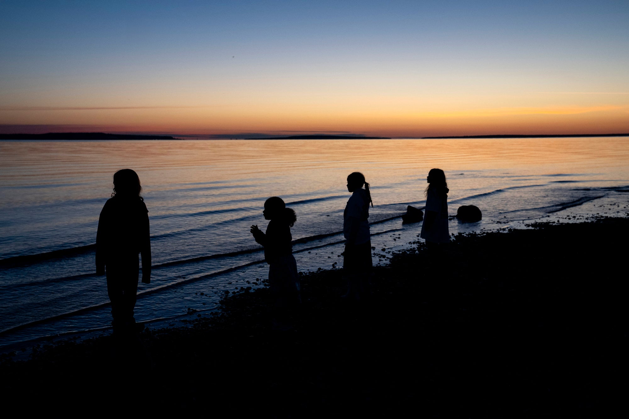 A silhouette of four children can be seen in front of lake water as a very early dawn brightens the sky before sunrise.