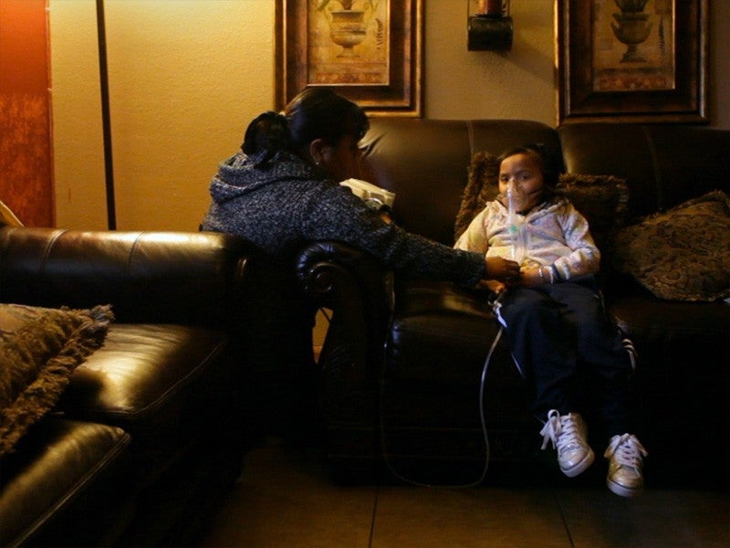 A child, seated in a reclining chair, receives treatment for asthma, as her mother kneels by her side and watches over here.