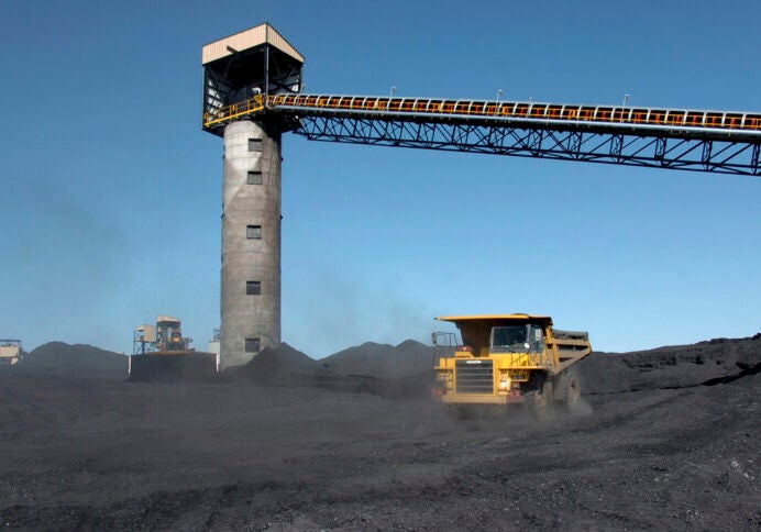 An industrial dump truck drives through a field of coal with some structures and towers in the background.