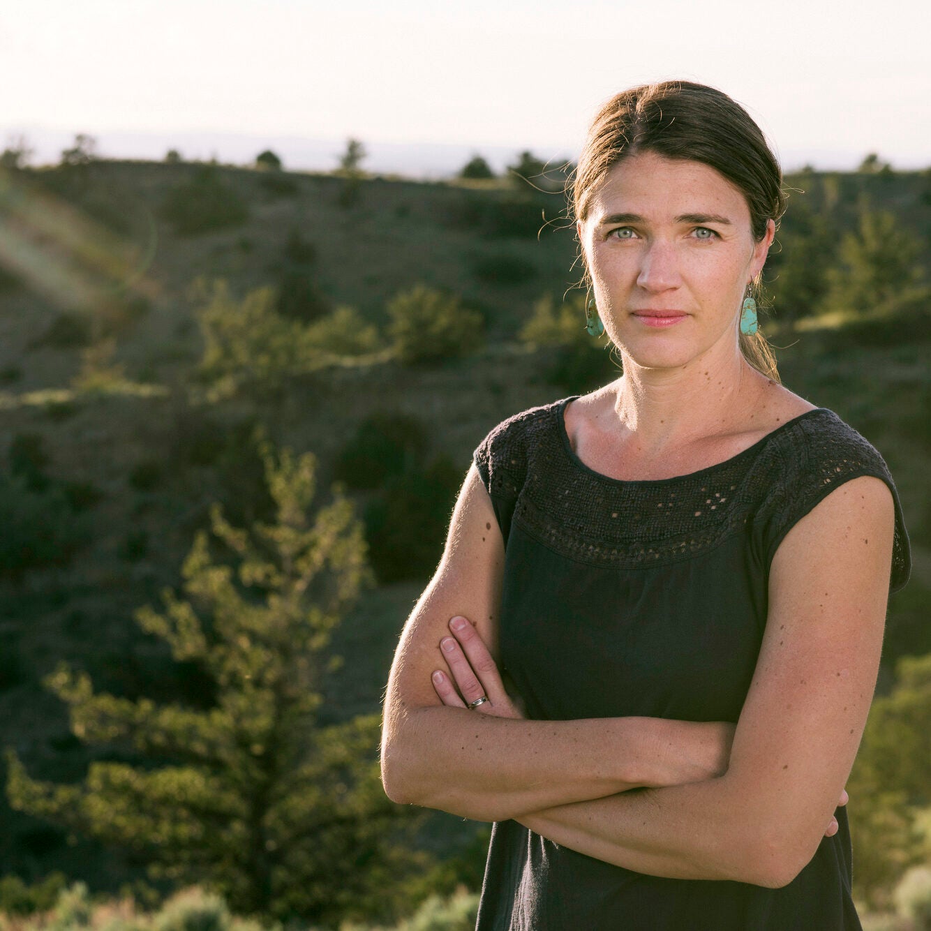 A woman stands with arms crossed outside with green hills and trees pictured behind her.