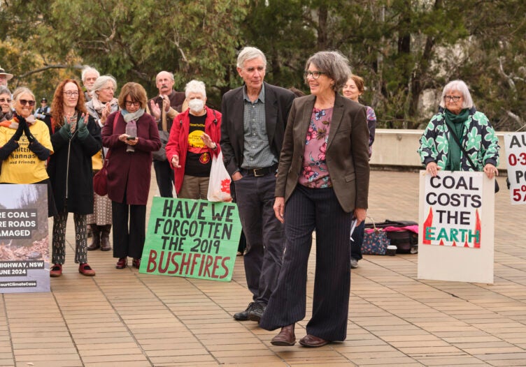 A group of people holding signs saying "Have we forgotten the 2019 bushfires" and "coal costs the Earth" applaud a man and woman in the foreground of the photo.