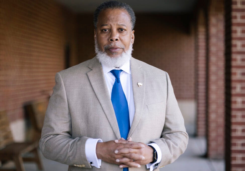 A man in a suit clasps his hands together and looks directly into the camera while posing for a portrait