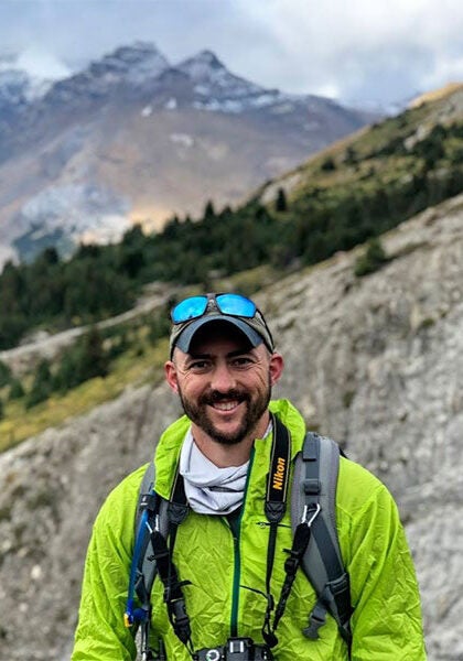 Aaron looks directly at the viewer with a friendly, open smile. He's outdoors, with a backpack and a camera hanging from his neck. The background, slightly blurred, is a sloped rockface, with thick green forests. In the distance is a snow-capped peak of another mountain.