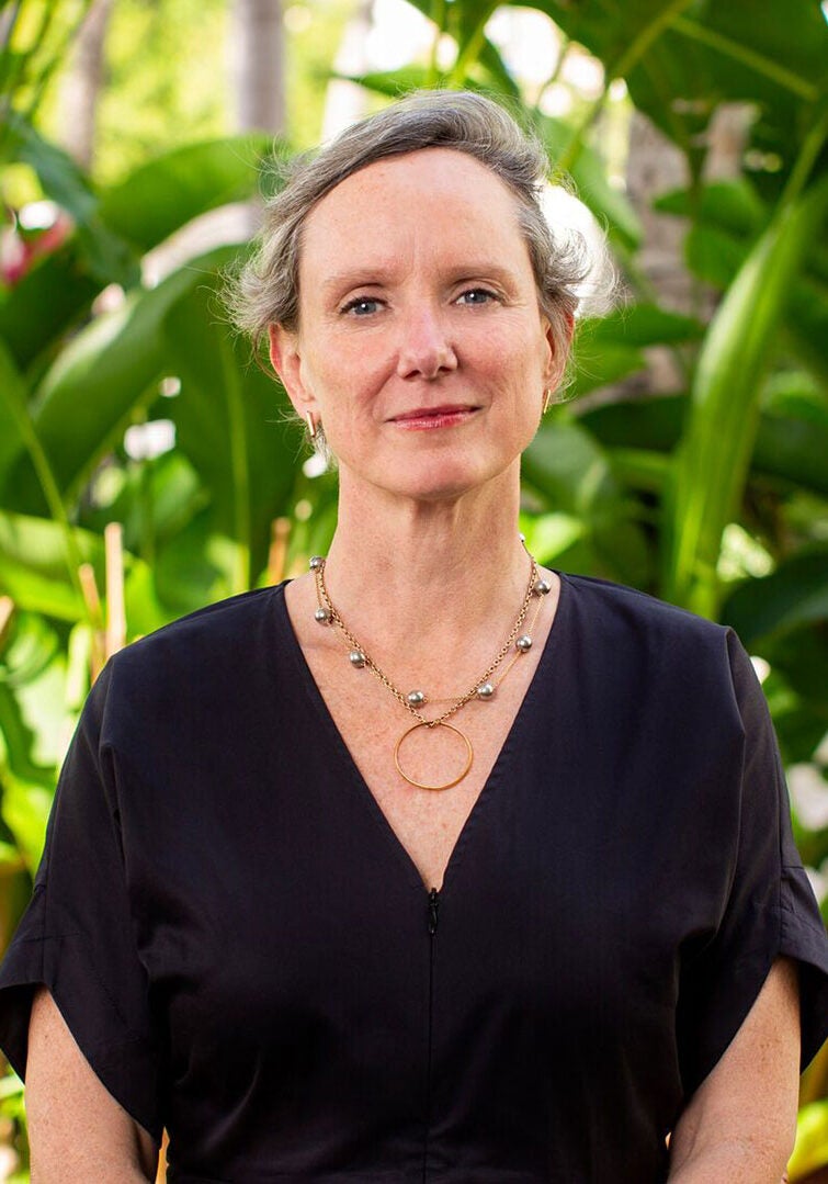 Abigail Dillen, President of Earthjustice, stands facing the viewer with a kind expression, in a brightly lit, verdant tropical landscape. She's wearing a black dress with simple pearl and gold circle necklackes.