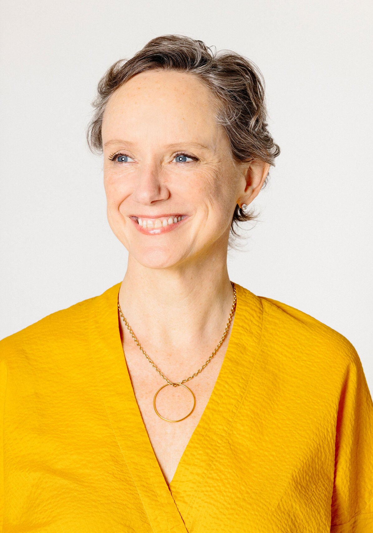 Abigail, with a friendly, open smile and bright, lively eyes, stands facing the viewer, looking slightly off to the viewer's left. She's wearing a bright yellow top and is standing in front of an off-white backdrop.