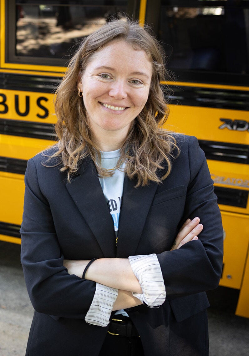 Ada stands facing the viewer with an open, friendly smile. She's wearing a black suit jacket, with the sleeves rolled up to her forearms showing the white with black striped pattern on the inside of the jack. Her arms are crossed and folded in front of her body. Ada's standing in front of a bright yellow school bus that runs on electricity. The day is sunny, with rays of sunshine reflecting off the windows of the bus.