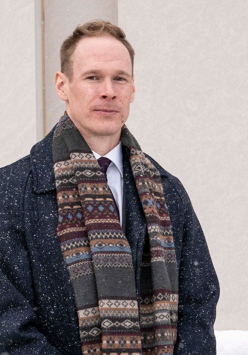 Adam looks directly at the viewer with a serious expression, as he stands outside in lightly falling snow, in front of a beige colored wall. He's wearing a white collar shirt and dark colored tie under a long black thick winter overcoat, with a brown and burgundy patterned scarf hangs loosely around his neck
