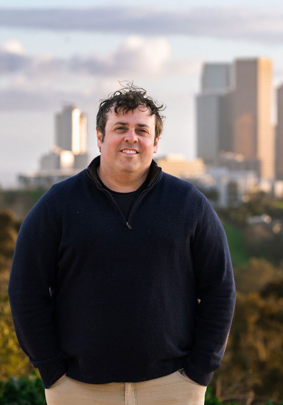 Adrian stands outdoors, wearing a dark-colored zip-up jacket and khaki slacks and looking directly at the viewer with a slight smile. His hands are in his pockets. The skyscrapers of the the Los Angeles skyline are visible behind him. The sun is shining on the buildings.