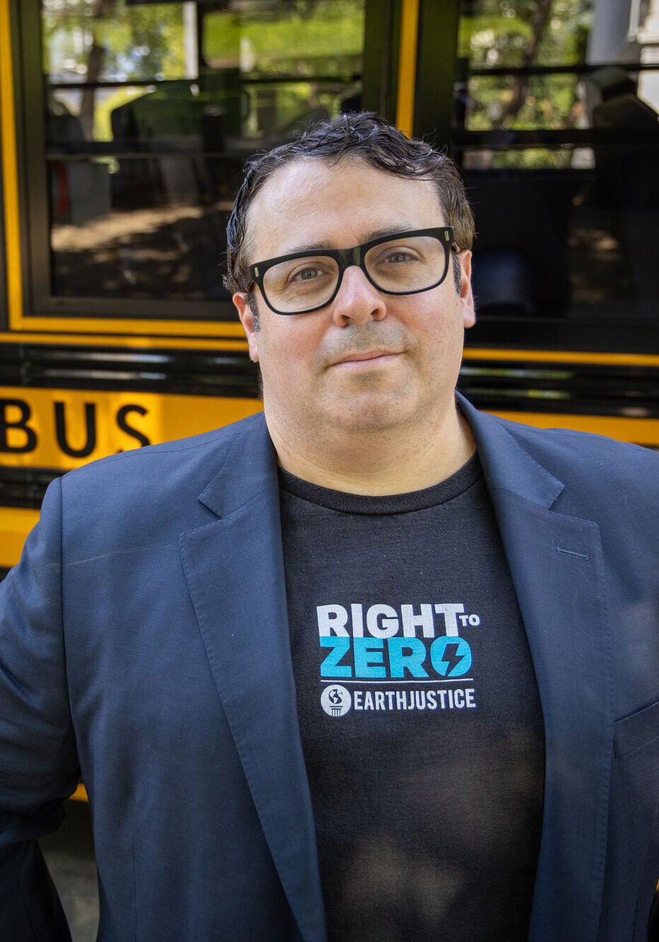 Adrian stands facing the viewer in front of a yellow electric school bus, with a slight smile. He's wearing a dark blue blazer jacket and black t-shirt with the Right to Zero and Earthjustice logos printed on it.