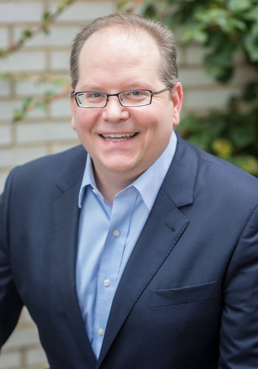 Andrew looks at the viewer with a friendly, open smile. He's wearing a dark blue suit jacket and a light blue collar shirt. He's in an outdoor setting. The background is blurred, with a brick wall and greenery visible.