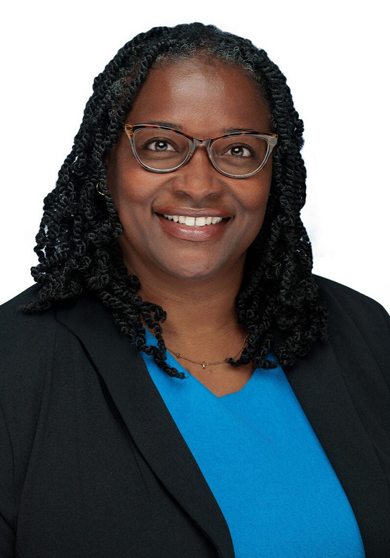 Anika looks directly at the viewer with a friendly, open smile. She's wearing a black suit jacket and a bright blue blouse. She's in front of a white backdrop.