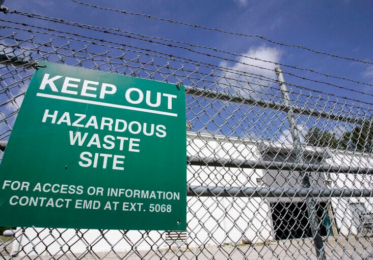 A green sign reading "Keep out hazardous waste site. For Access or information contact EMD at Ext. 5068" hangs on a chain link fence in front of a white cinder block building.