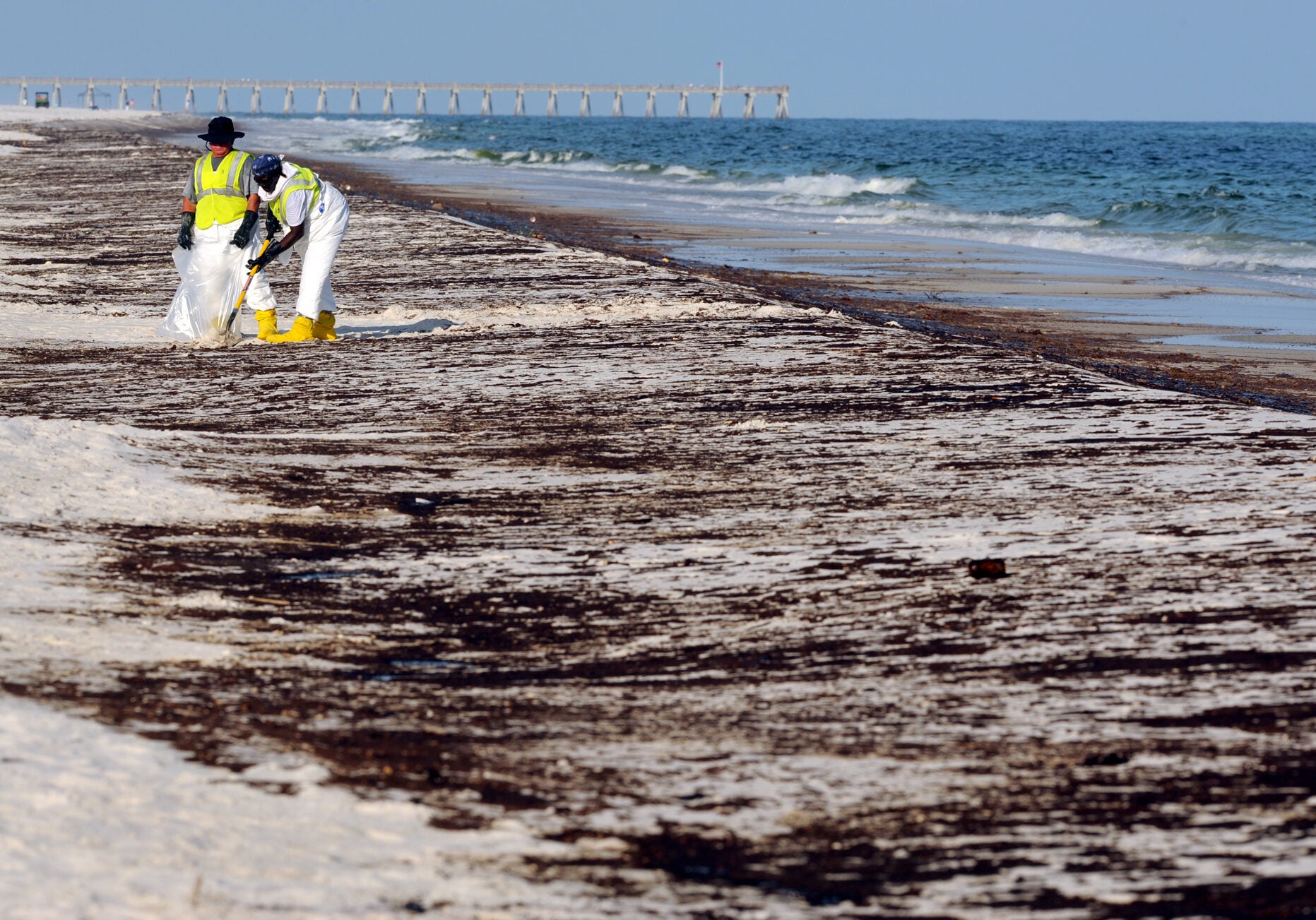 Two people in hazmat suits and work vests scoop oil covered sand into a bag on a mostly empty oil covered beach. The are smaller in the picture and there are a few people in the far distance.