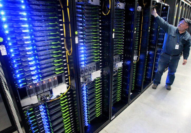 Racks of servers with blue LED lights sit in a room as a man works with something in the background.