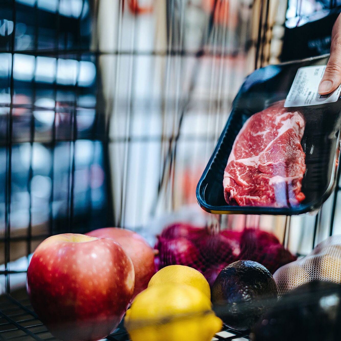 A person places a plastic-wrapped container of raw beef into a shopping cart, which already holds apples, lemons, avocadoes, and other produce.