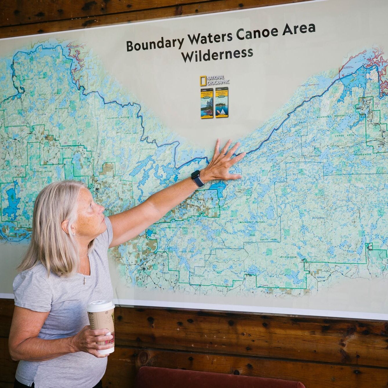 Becky Rom, wearing a light grey t-shirt and holding a large to-go coffee cup with a lid on it in her right hand, is reaching up and stretching out her left hand against an extremely large map of the Boundary Waters wilderness that's mounted on a wood-planked wall. She's looking up to where her left hand's fingers are outstretched. The map shows that the wilderness area borders the northern boundary of Minnesota with Canada.
