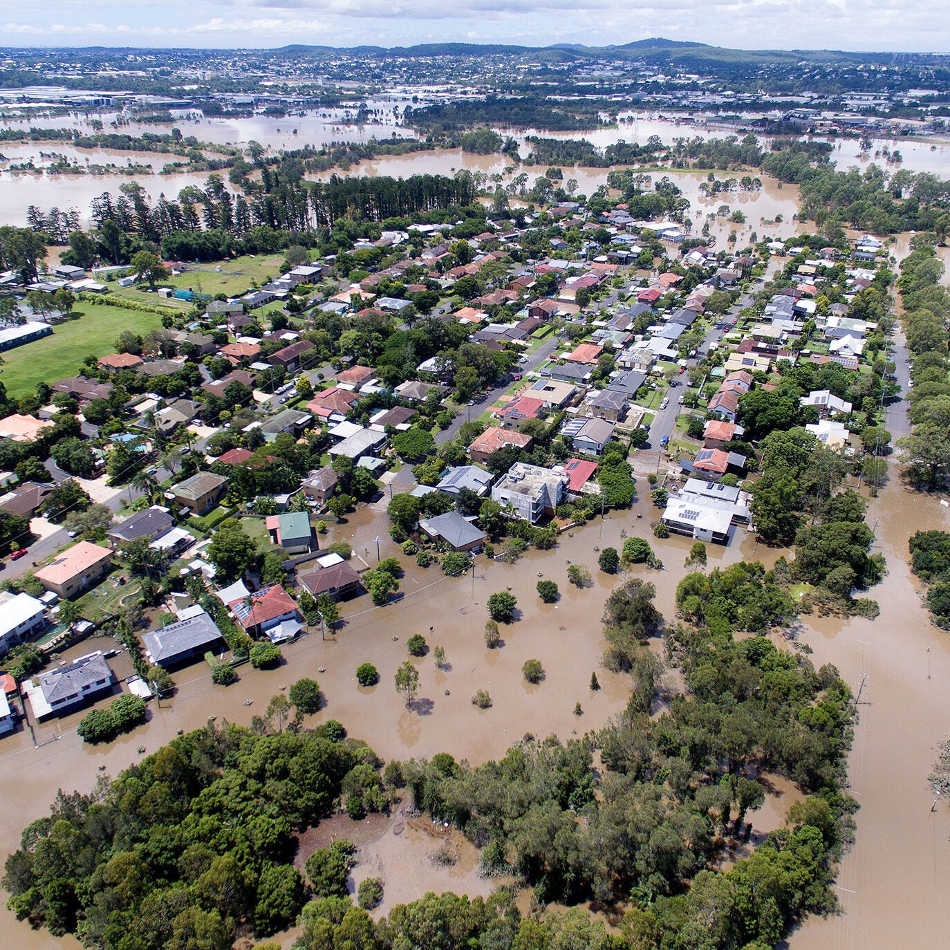 Aerial view of dozens, perhaps hundreds, of homes, partially submerged in murky, brown water. The floodwaters seem to stretch for miles. Bright green trees are visible above the floodwaters.