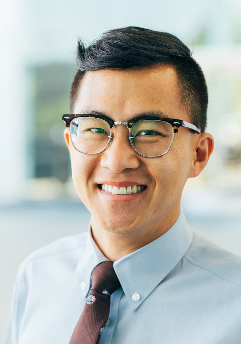 Byron looks directly at the viewer with a friendly, open smile. He's wearing a light blue collared shirt and a dark brown tie. The background is blurred and appears to be an interior, bright, airy space.