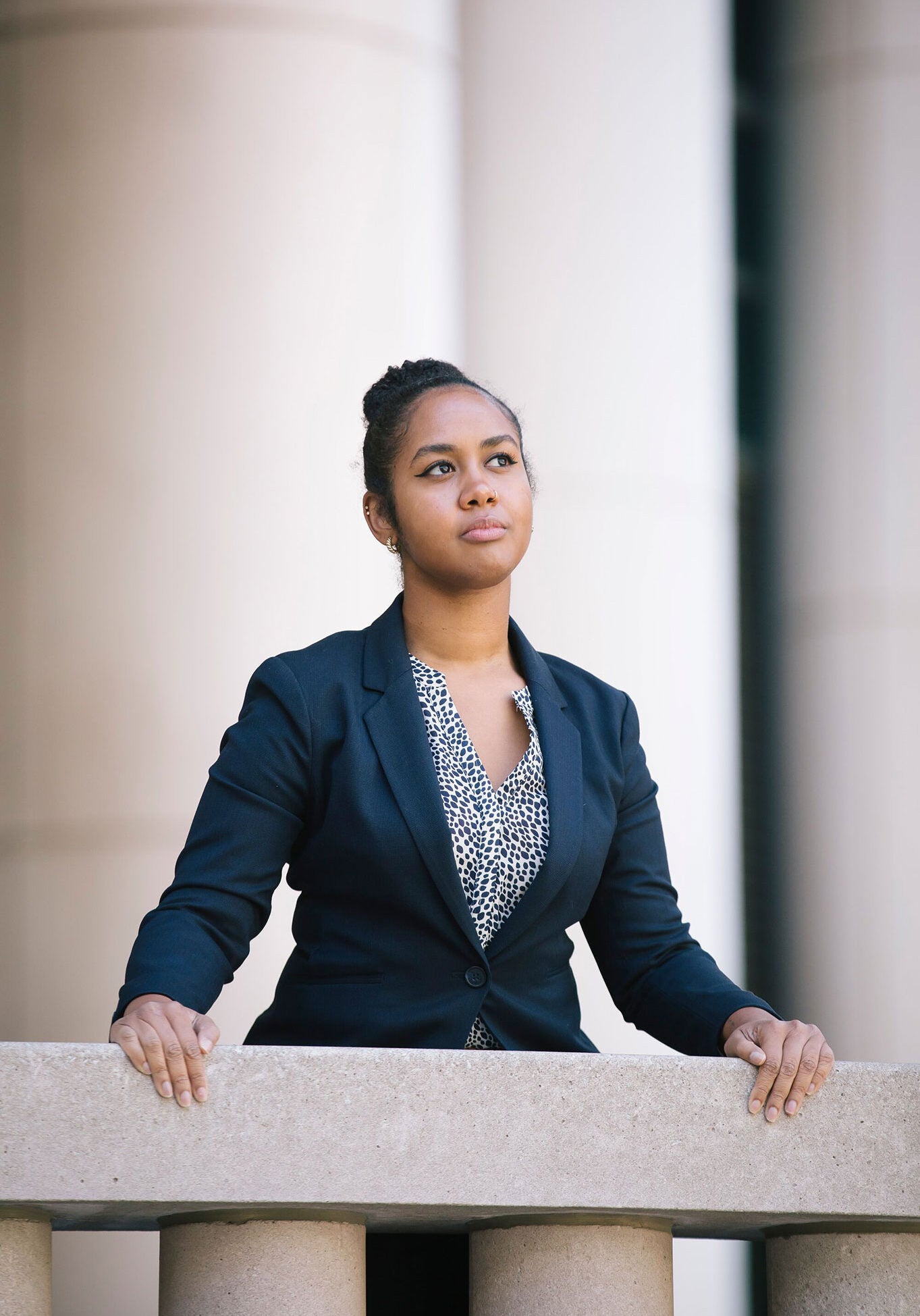 Candice stands with her hands resting on a stone railing in front of her, outside of a building with large stone columns. She is wearing a black suit and a white and black patterned blouse. Candice looks slightly upwards with a contemplative expression. The background is blurred and out of focus.
