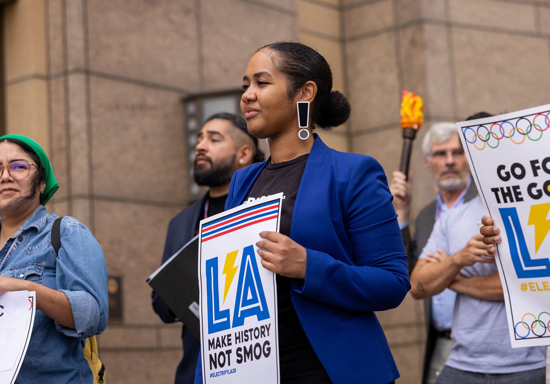 A group of people stand together holding signs that say &quot;LA: Make history not smog&quot; and &quot;Go for the Gold, LA. #ElectrifyLA28.&quot;