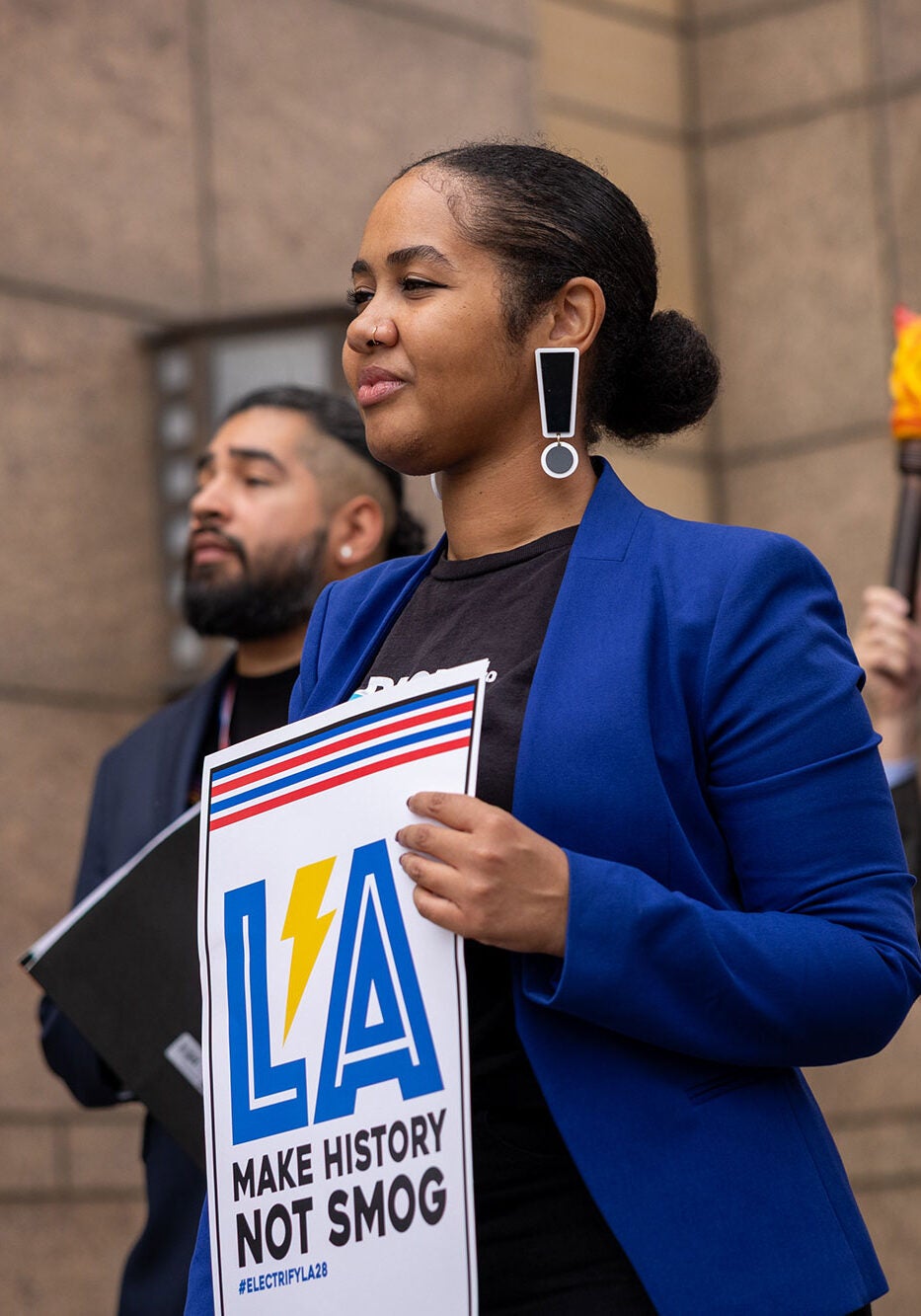 A group of people stand together holding signs that say &quot;LA: Make history not smog&quot; and &quot;Go for the Gold, LA. #ElectrifyLA28.&quot;