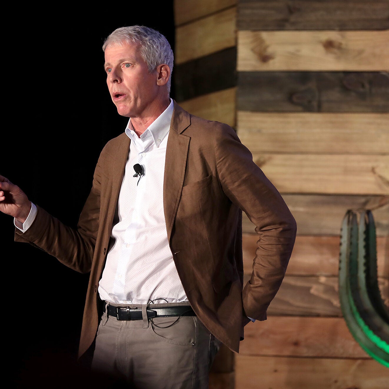 A man wearing a white shirt and brown jacket speaks on a stage with a cactus.