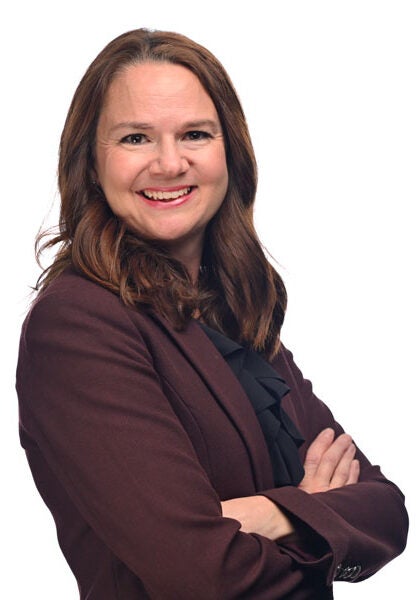 Christie, wearing a burgundy suit with a black blouse, stands in front of a white backdrop. Her body is angled to the viewer's right, and her arms are folded in front of her chest in a firm, but relaxed way. She looks over her right shoulder to the viewer with a friendly, wide smile.
