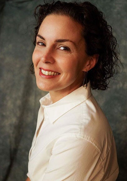 Conchita looks at the viewer with a friendly, open smile. Her body is angled to the left, and her head is turned towards the viewer. She's wearing a white, collared shirt, and stands in front of a grey backdrop.