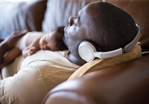 A father and newborn baby daughter at home sleeping. He is wearing headphones and wearing casual clothing.