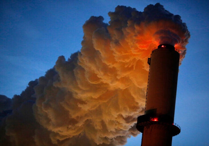 Dark gray clouds of steam emerge from the top of a smokestack at night, lit by red lights on the smokestack with a dark blue sky in the background.