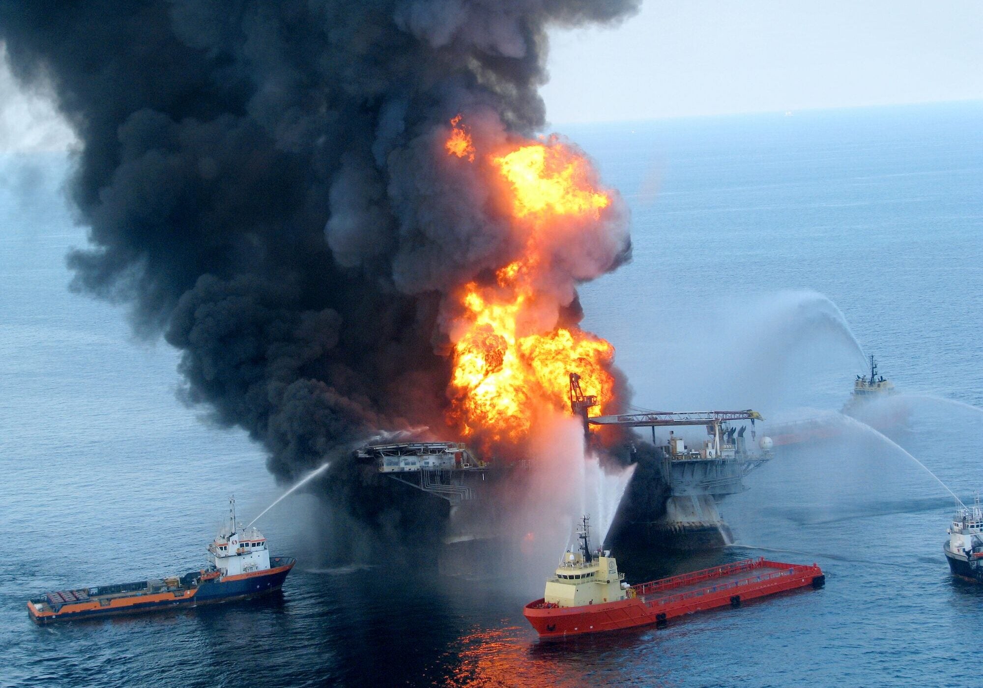 Large flames and smoke come out of an offshore oil platform as several boats spray water onto it in the ocean.