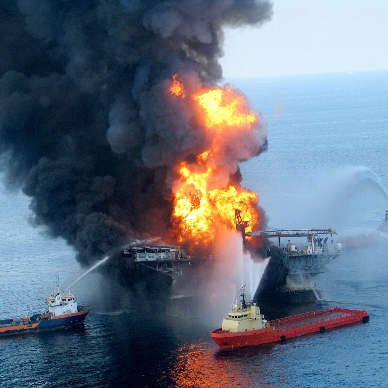 Large flames and smoke come out of an offshore oil platform as several boats spray water onto it in the ocean.