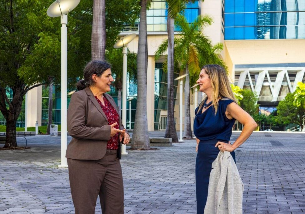 Two women stand outside of a large building surrounded by trees talking.
