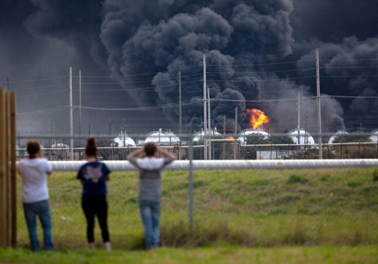 People standing in front of a fence viewing a fire consume a chemical plant
