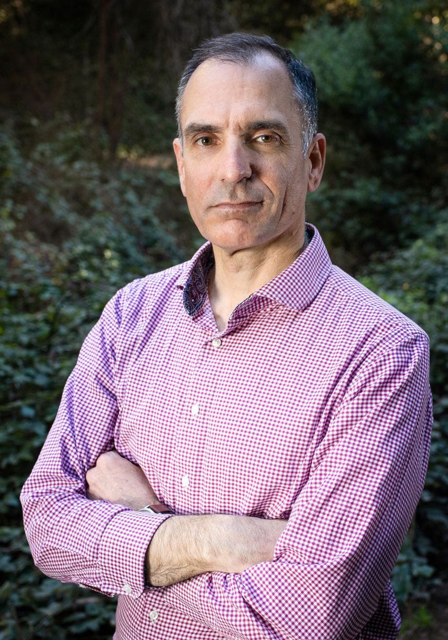 Drew looks directly at the viewer with a serious expression. His arms are crossed in front of him, and his body is slightly angled to the viewer's left. He's in a verdant, outdoor area. The greenery in the background is blurred. Drew is wearing a red and white checkered collared buttoned down shirt.