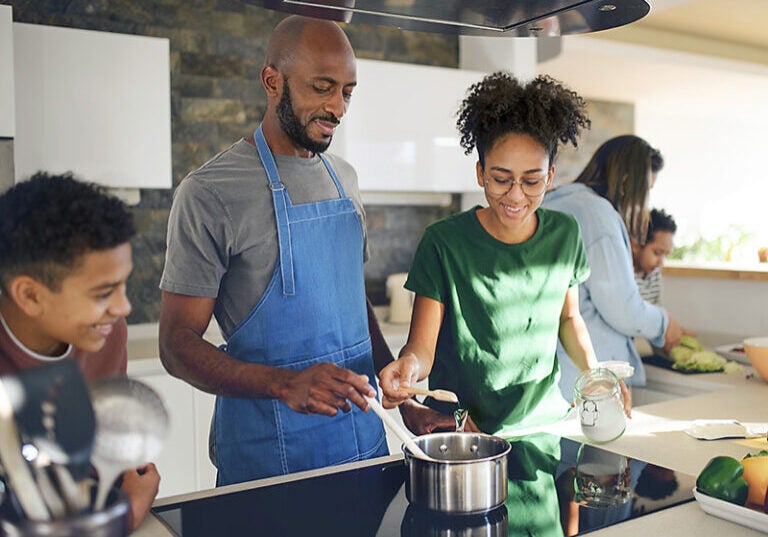 A man wearing a grey shirt and blue apron stands in front of an electric range top. With one hand, he is holding a white utensil to a steel saucepan. To his left (our right) is a young woman in glasses and a green t shirt. She is holding a jar of something white in one hand and with her other, she is pouring what she scooped out of the jar into the pan. To the man's right (our left) is a young man leaning on the counter beside the stove, smiling at the saucepan. In the background are two other people looking at a chopping board as if prepping ingredients.