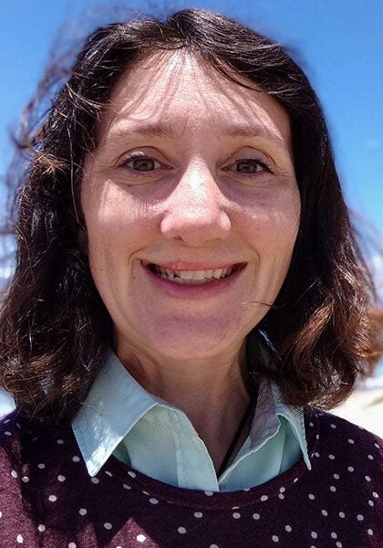 Elizabeth looks directly at the viewer with a friendly open smile. She's wearing a white collar shirt under a burgundy patterned sweater. Elizabeth is standing on a sunny beach, with the wind gently blowing and the sun shining down. The sky is a brilliant, cloudless blue.