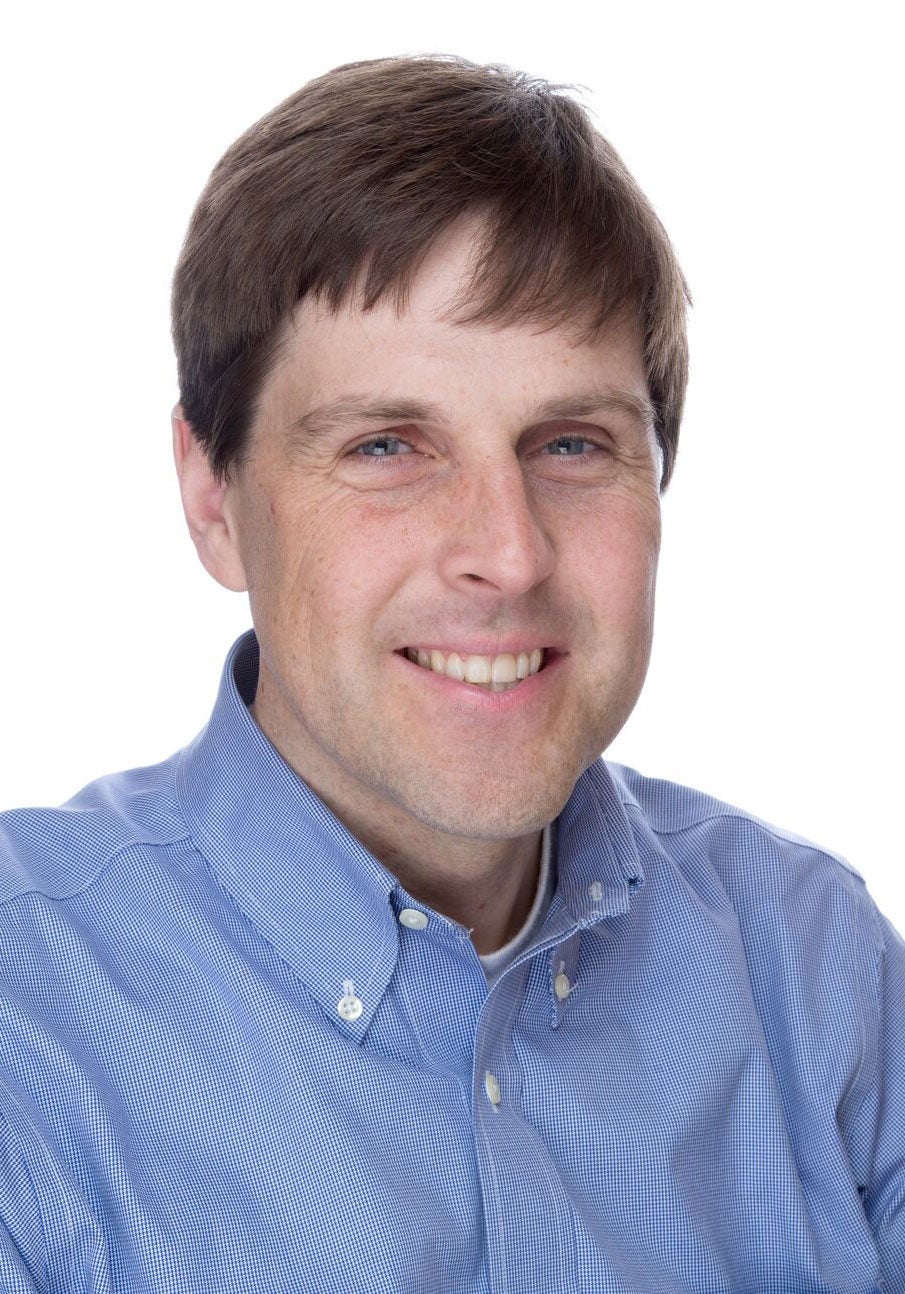 Erik looks directly at the viewer with a friendly, open smile. He's wearing a collared button-down shirt with a blue and white checkerboard pattern. He's in front of a white backdrop.