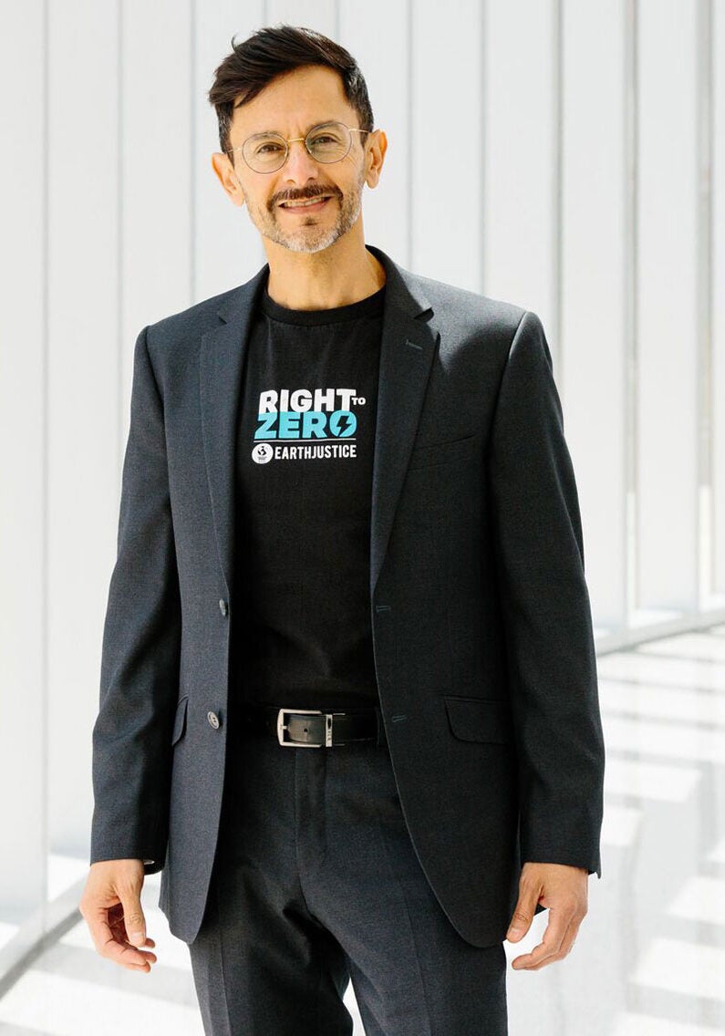 Fernando stands looking directly at the viewer with a friendly smile. He is wearing an all-black suit with a black t-shirt that has the logo for Earthjustice's Right To Zero campaign on the front. In the logo, the word "Zero" is in bright blue, with the rest of the text in white. Fernando is standing in an all-white stairwell, where the walls are slim vertical white pillars.