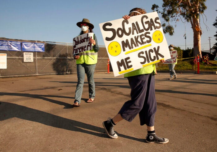 People hold signs in front of a gated facility. One says "SoCalGas makes me sick"
