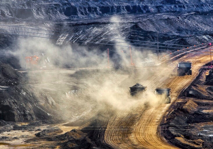 A photo showing black, terraced earth with a dirt road going through it. There are three dump trucks driving on the road kicking up a lot of dust.