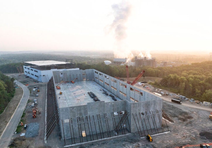 Two large buildings under construction in morning light, with a power plant in the background with steam coming out.