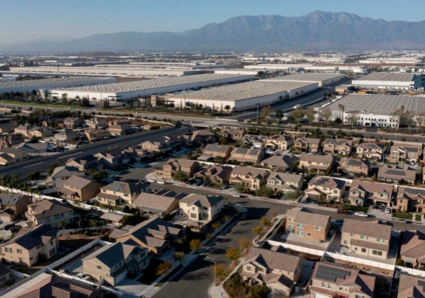 Suburban houses in the foreground are next to an expanse of large, white warehouses that stretch into the distance. The horizon is hazy with mountains in the distance.