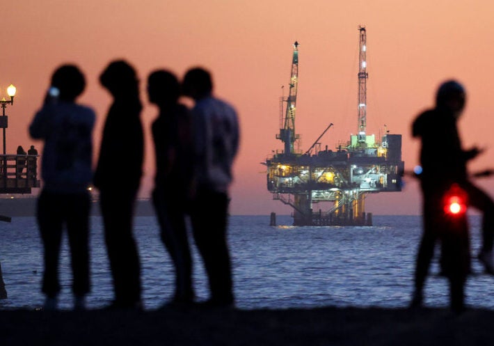 A group of silhouetted people stand on the shore in the evening with a lit up drilling platform visible in the distance.