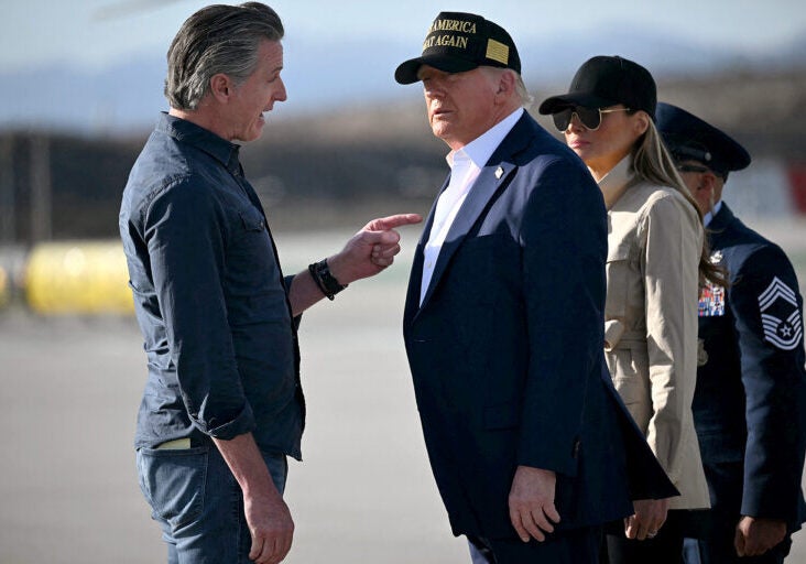 Gavin Newsom, wearing jeans and a blue collard shirt is pointing a finger at Donald Trump's chest while speaking to him with a serious expression. Trump, wearing a blue suit jacket and no tie and a black baseball hat, is looking at him.