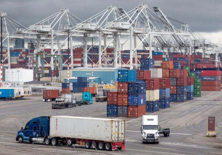 Semi trucks drive through stacks of metal containers in a busy shipping port. Large cranes for removing containers from ships are in the background.