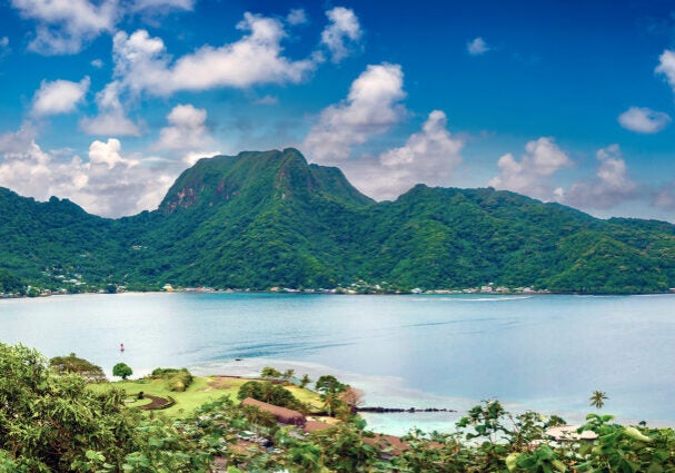 A wide landscape view of a green island wrapping around a large, natural, blue ocean harbor. Small buildings are on the coast with a tall hill or mountain on the far side.