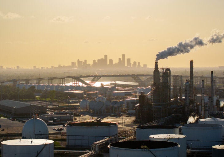 A refinery with storage tanks and exhaust stacks, some releasing vapor, are in the foreground of an aerial photo with the skyline of Houston on the horizon.